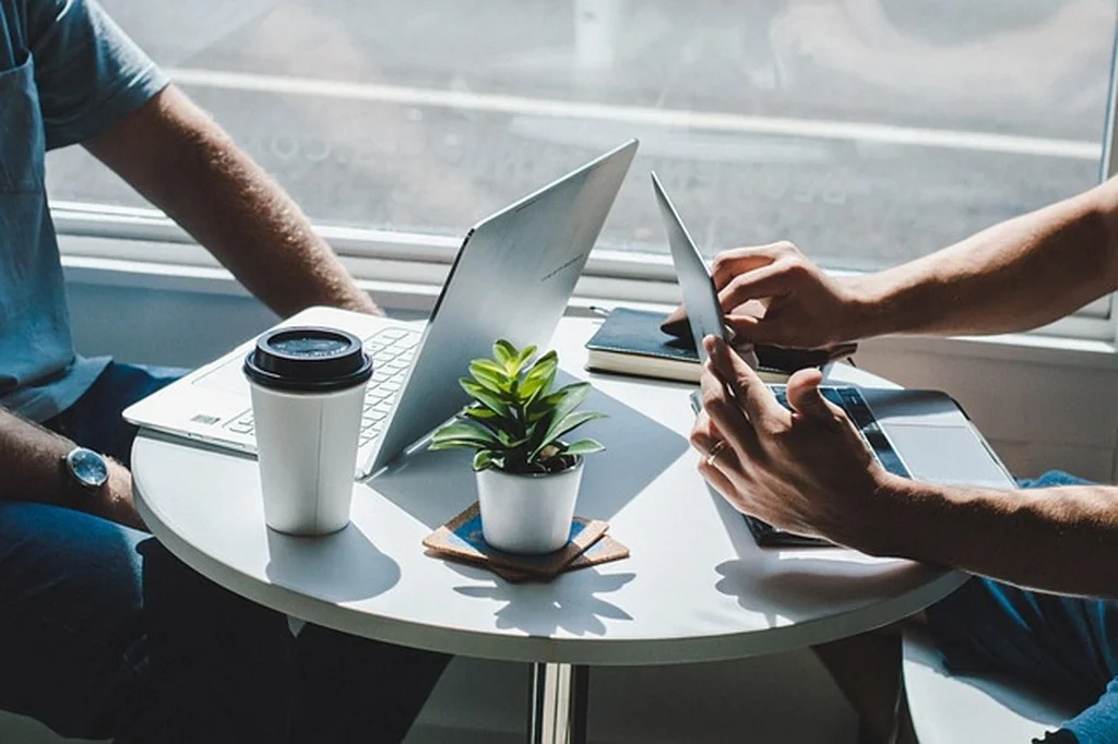 Duas pessoas à mesa num ambiente comercial. Sobre a mesa está um pequeno vaso de plantas, um copo de café com tampa, um caderno de anotações, e dispositivos como notebook e tablet. As pessoas simbolizam estar fazendo controle de aluguel via software de controle