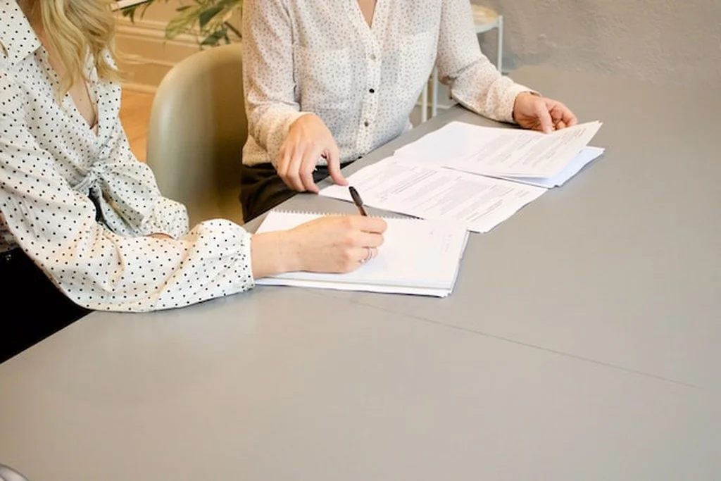 Contratos sobre a mesa, uma mulher arrumando alguns deles e a outra assinando um deles, simbolizando o distrato imobiliário