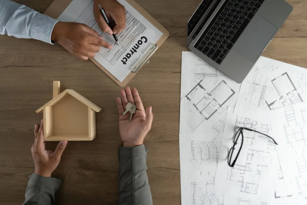 Uma pessoa assinando um contrato e a outra entregando chave de imóvel, enquanto segura uma casa de madeira, simbolizando um negócio feito em plantão de vendas. Além do contrato e da casa de madeira, sobre a mesa também está um notebook, o desenho da planta de uma casa e óculos de grau sobre o projeto/desenho