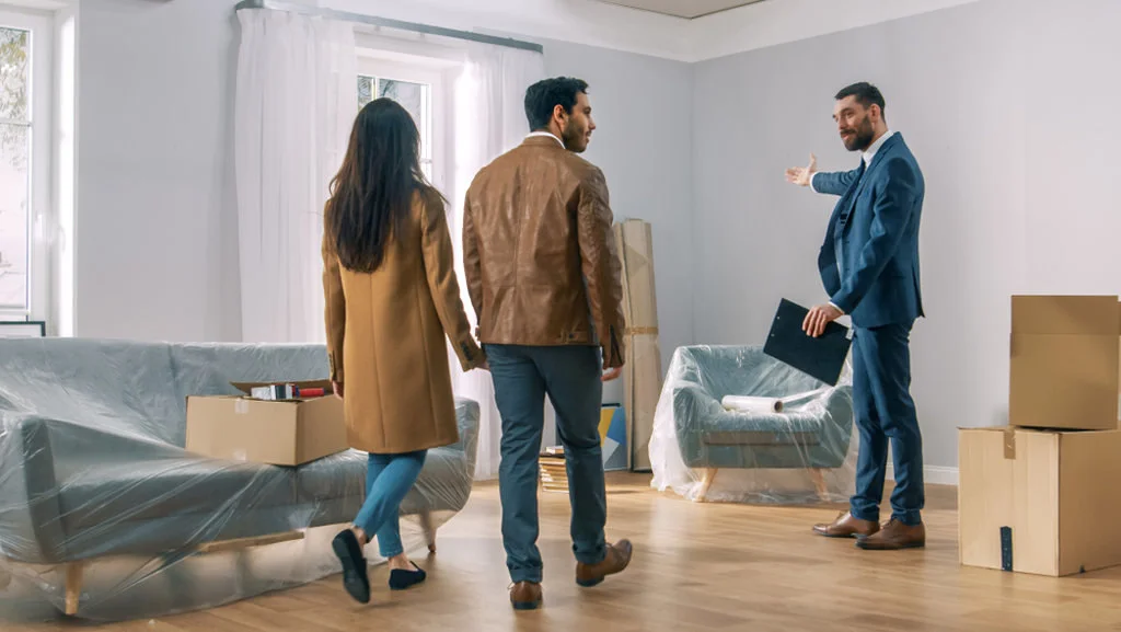 Corretor de imóveis mostrando para um casal uma sala de estar com móveis cobertos/protegidos. Simbolizando quem avalia a precificação de imóveis
